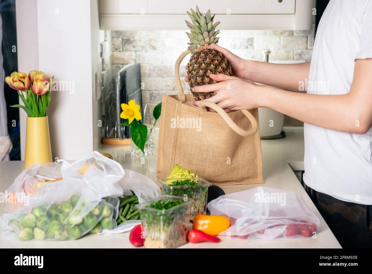 Mann, der Ananas aus wiederverwendbaren Stoffbeuteln mit Gemüse auf dem weißen Küchentisch nimmt. Frisches Bio-Gemüse. Online-Essen zu Hause Stockfoto
