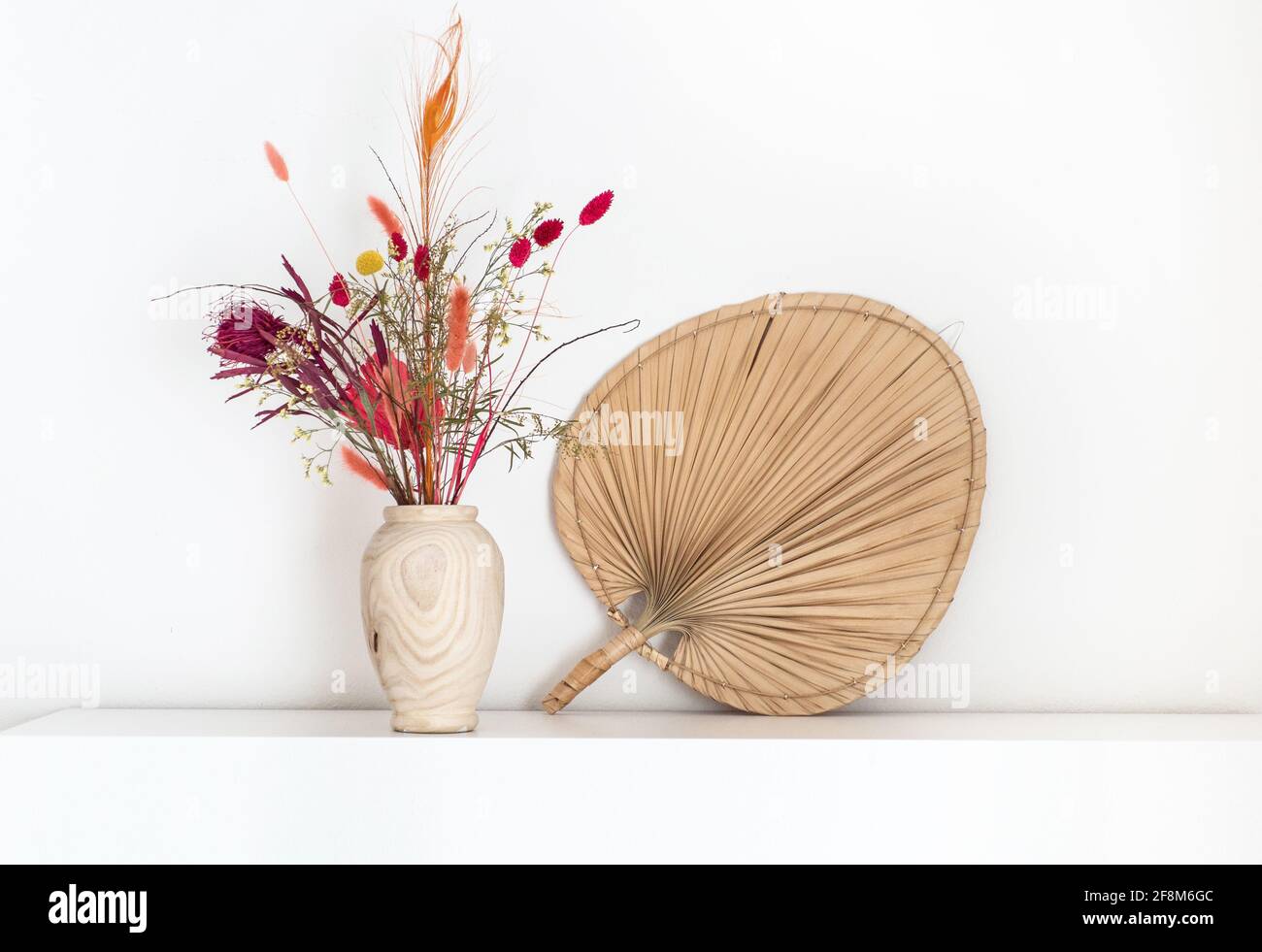 Buntes getrocknetes Blumenstrauß in einer hölzernen Vase im Boho-Stil, die auf einem weißen Regal mit einem Fächer aus getrocknetem Pam-Blatt daneben steht. Stockfoto
