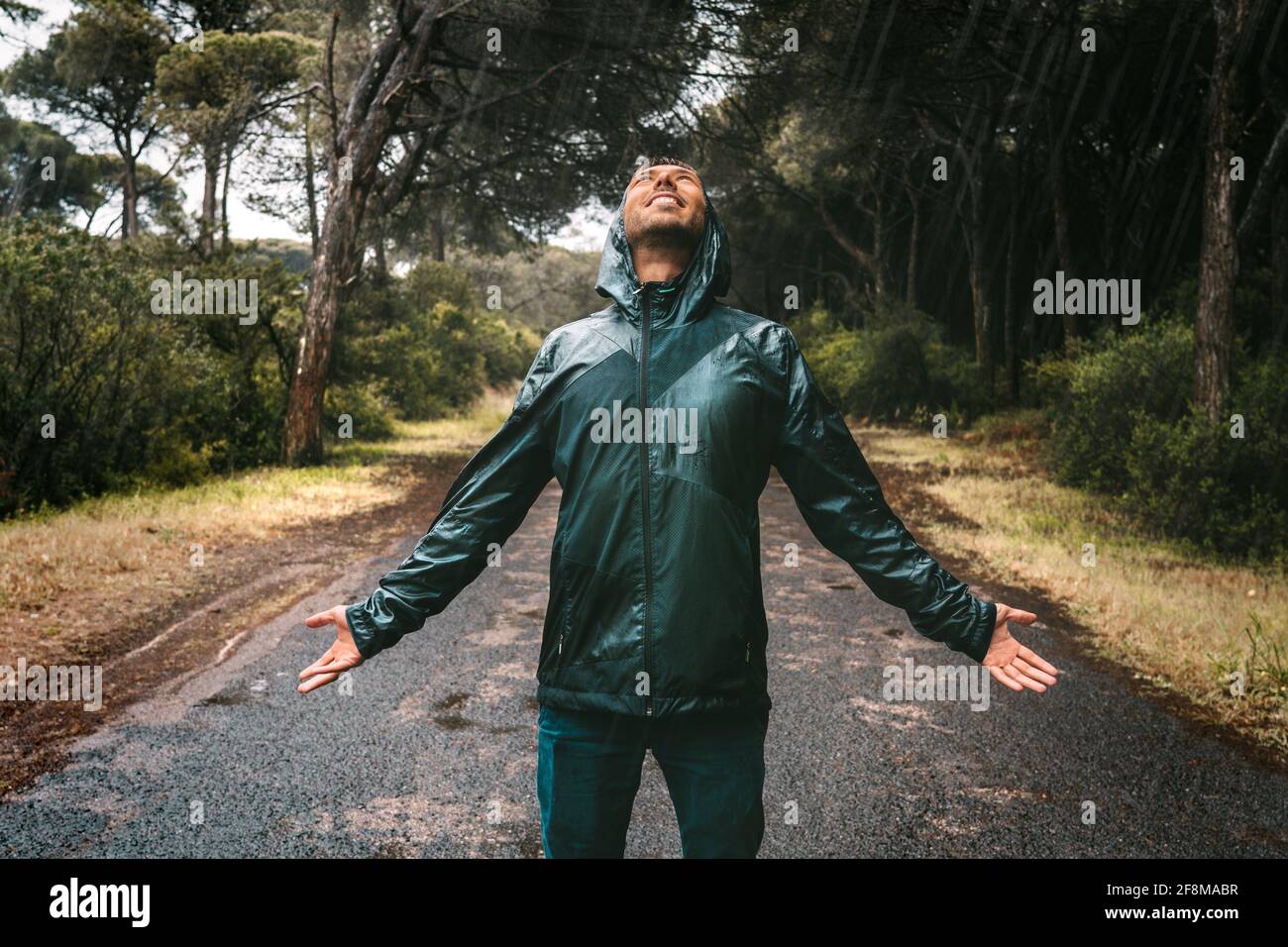 Junger Mann mit nassem Lächeln in einer durchnässten Jacke mit Kapuze, der den Regen genießt. Mann im Regenmantel stand bei starkem Regen. Schlechter Herbst, egal. Stockfoto