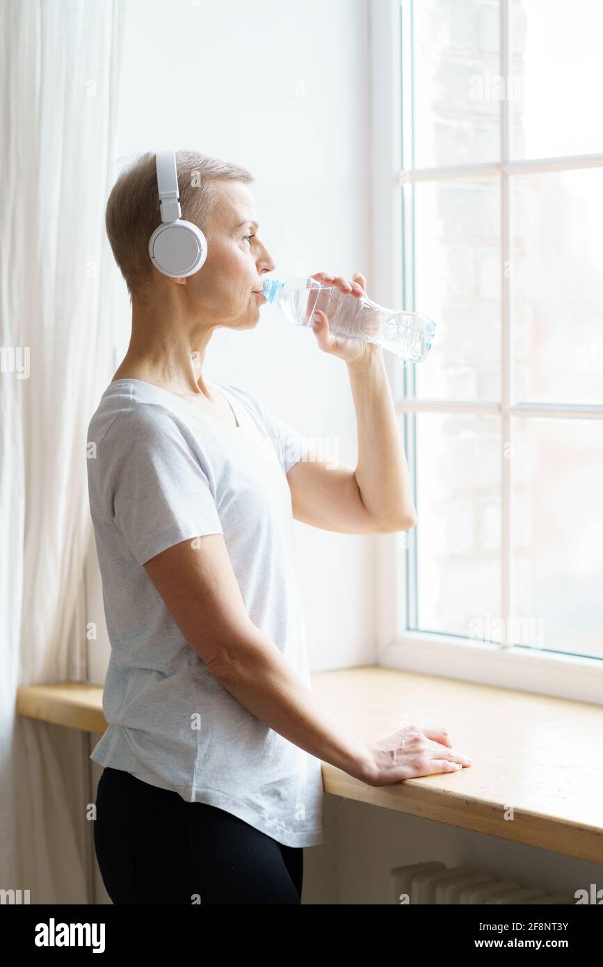 Vertikales Waistup-Porträt einer glücklichen älteren Frau, die nach der Fitness zu Hause Wasser aus der Plastikflasche trinkt. Reife Weibchen löschen Durst nach Wasser, das zum Fenster schaut. Ältere Frau nach dem Sporttraining Stockfoto