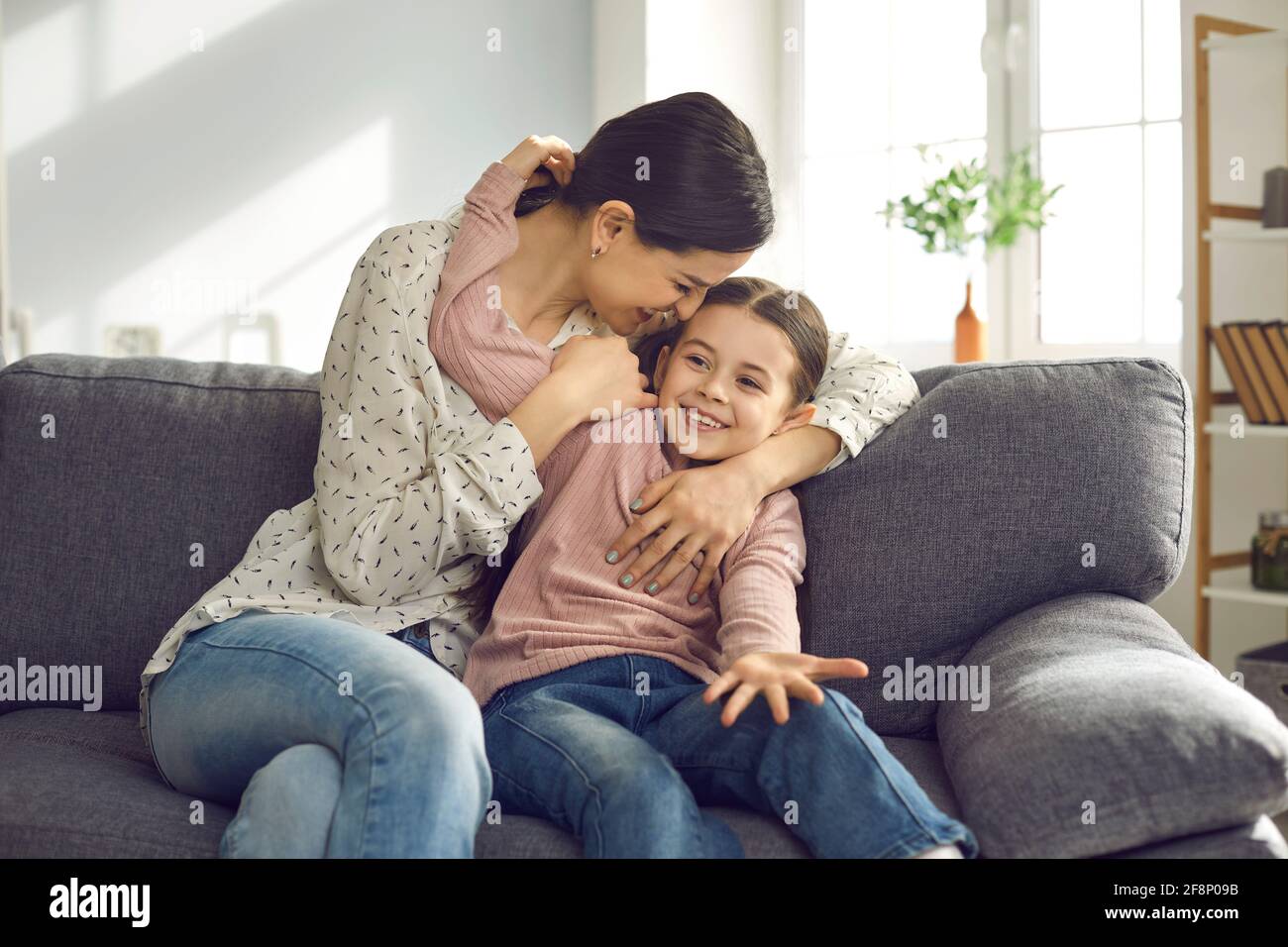 Fröhliche junge Mutter und Tochter haben Spaß, spielen und umarmen, während sie zu Hause auf der Couch sitzen. Stockfoto