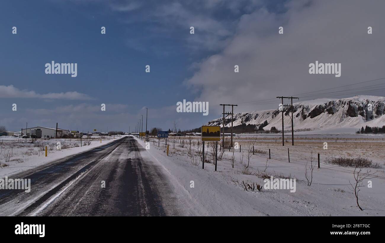 Blick auf den Dorfeingang der kleinen Stadt Kirkjubaejarklaustur im Süden Islands am sonnigen Wintertag mit eisbedeckter Straße und Ortsschild. Stockfoto