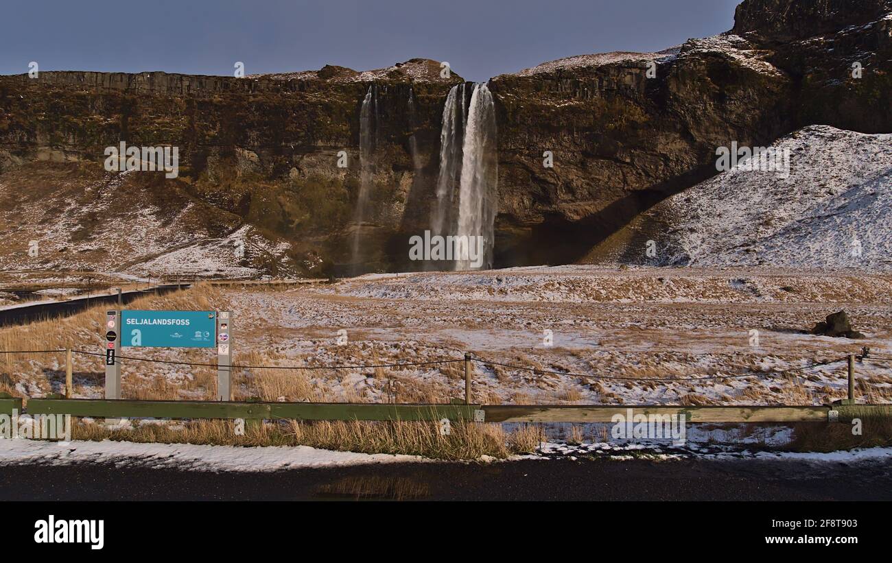 Schöne Vorderansicht des berühmten Wasserfalls Seljalandsfoss in der Wintersaison mit schneebedeckter Wiese und Namensschild. Stockfoto