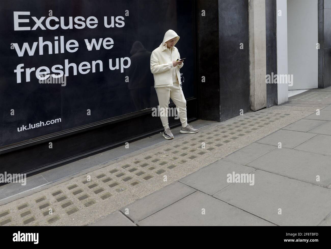London, England, Großbritannien. Ein junger Mann, der in Creme gekleidet ist und auf diesem Handy vor einem ÜPPIGEN Laden aussieht Stockfoto