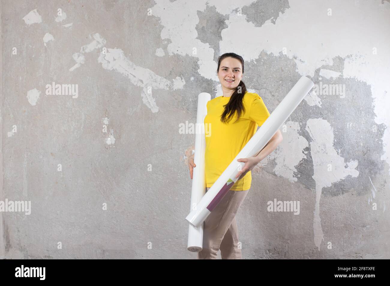 Eine Frau in einem leuchtend gelben T-Shirt hält 2 Tapetenrollen gegen die alte Wand. Reparaturarbeiten, Tapezieren. Mit einem Platz für Text, um Platz zu kopieren. Stockfoto