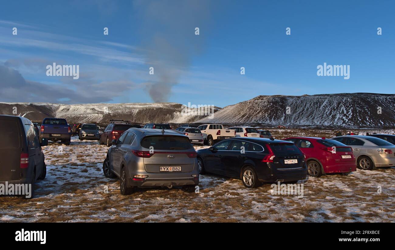 Autos parken auf der Wiese am Ausgangspunkt des Wanderweges zum Vulkanausbruch am Fagradalsfjall mit dem Rauch des Vulkans. Stockfoto
