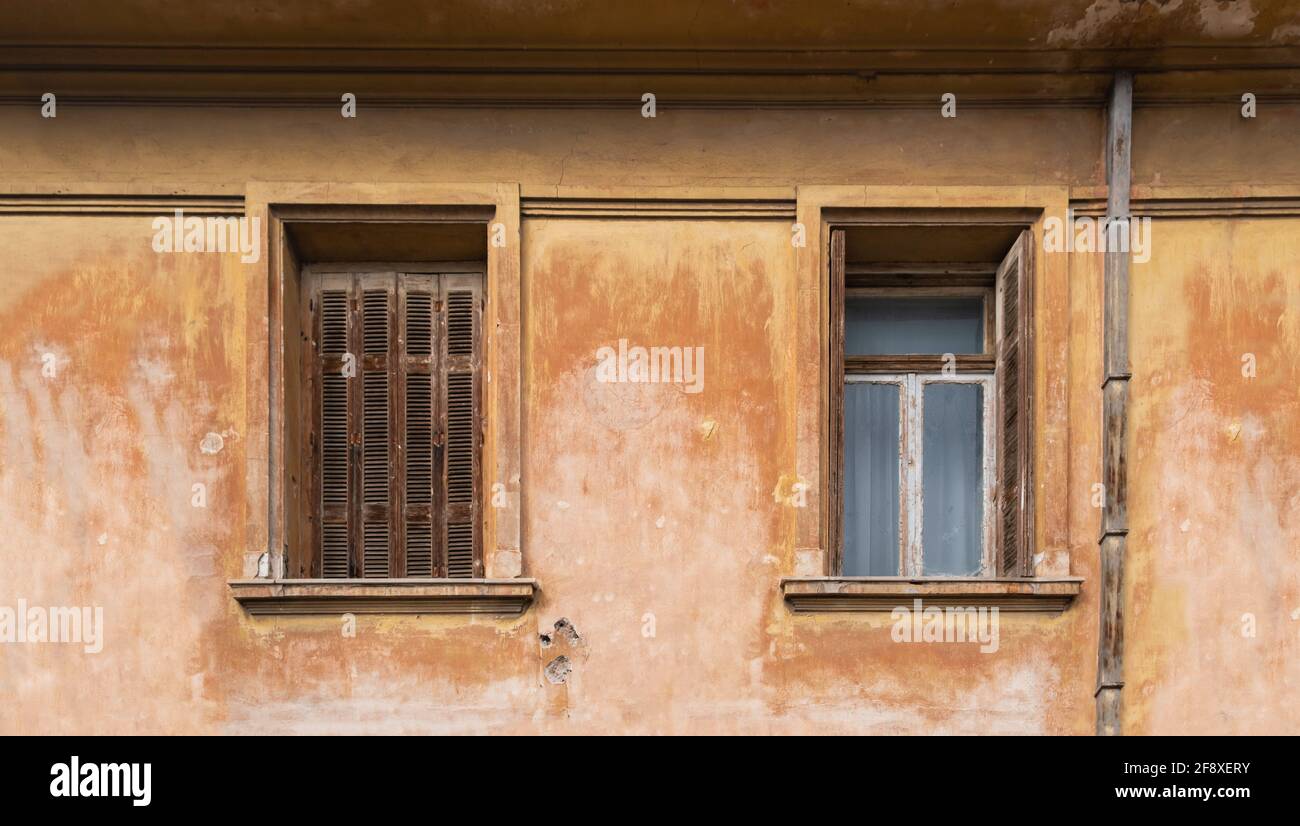 Verlassene Fenster mit offenen und geschlossenen Fensterläden an der Fassade eines verlassenen Wohngebäudes. Zwei Fenster auf schmutzigen braunen Wandhintergrund. Stockfoto