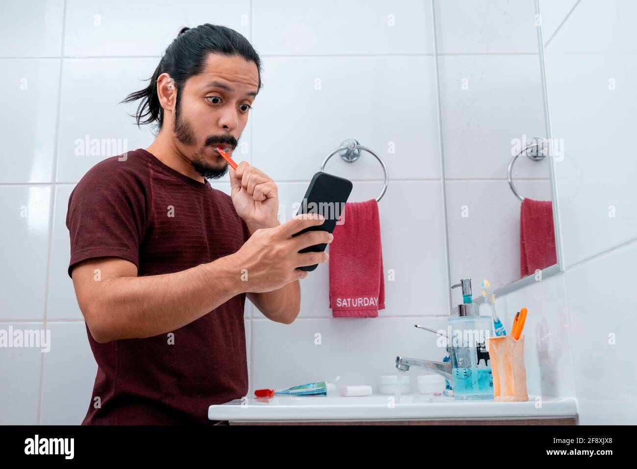 Junger latein mit langen schwarzen Haaren, der das Telefon benutzt Beim Zähneputzen im Badezimmer Stockfoto