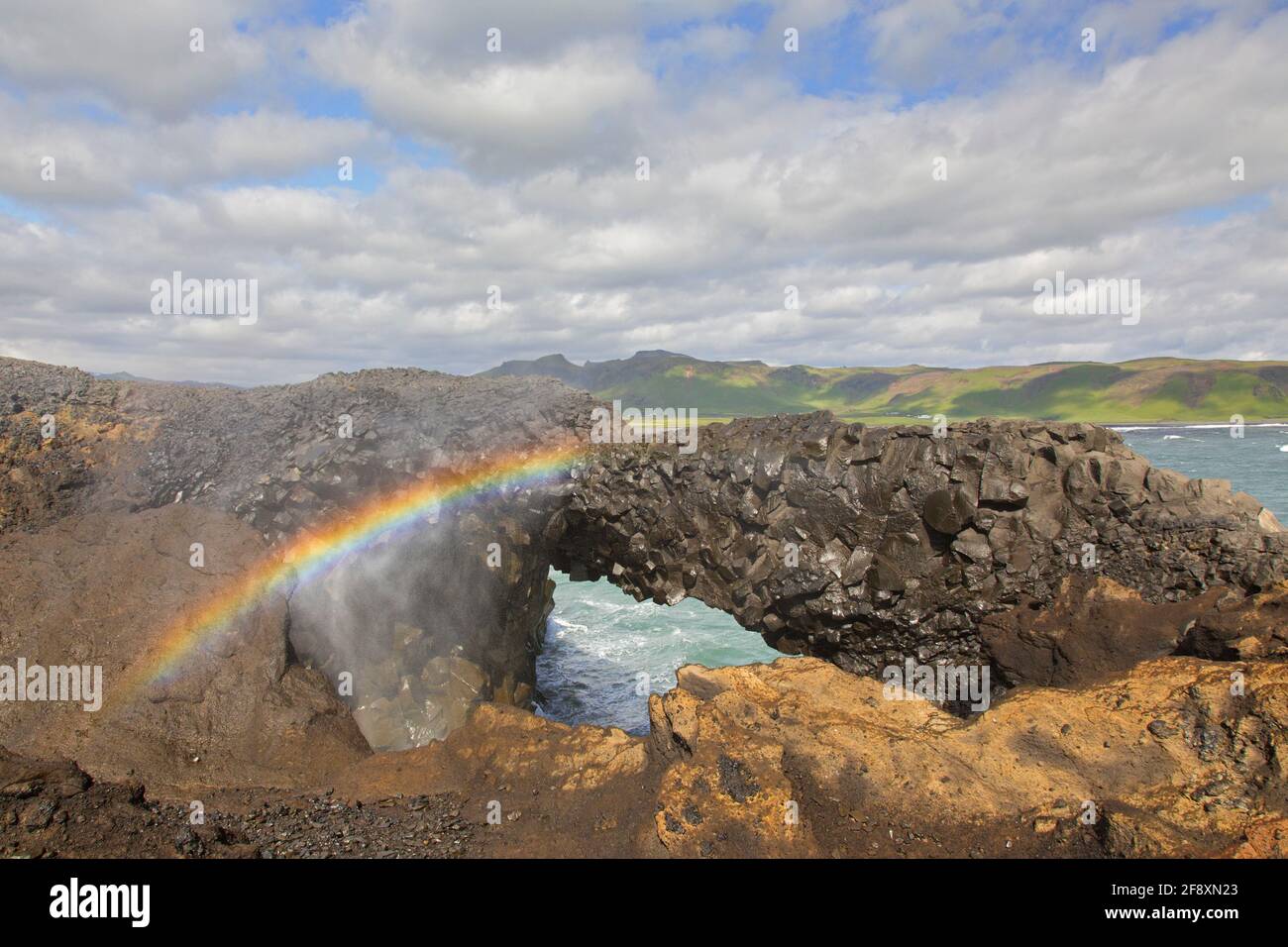 Regenbogen und natürlicher Bogen, erodierte schwarze Basaltsteinformation am Kap Dyrhólaey / Kap Portland bei Vík í Mýrdal, Island Stockfoto