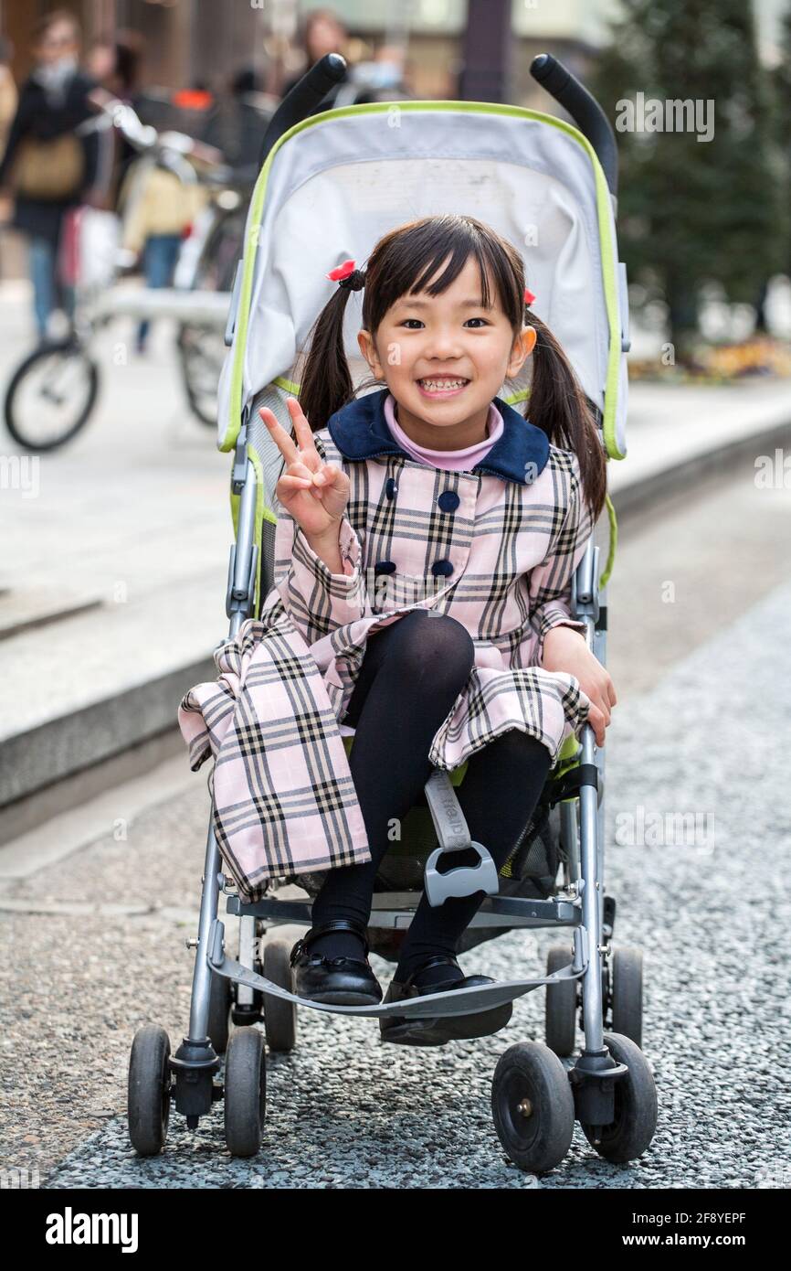 Nettes junges, wohlhabendes japanisches Kind gibt ein Friedensschild, das im Kinderwagen der jüngeren Schwester, Ginza, Tokio, Japan, sitzt Stockfoto