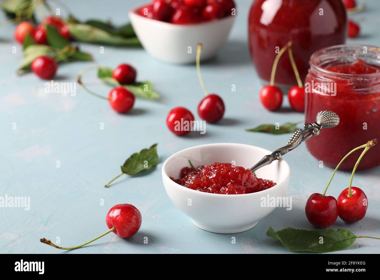 Hausgemachte Süßkirschkonfitüre in einer weißen Schale und Gläser auf hellblauem Hintergrund. Nahaufnahme Stockfoto