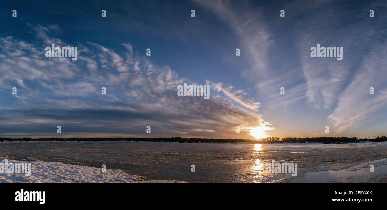 Landschaftlich weites Panorama des Frühlings-Sonnenuntergangs über dem schmelzenden eisigen See. Wunderschöne rot-gelb-orange Sonne, dunkelblaue und weiße Wolken am blauen Himmel. Nördliches Scand Stockfoto