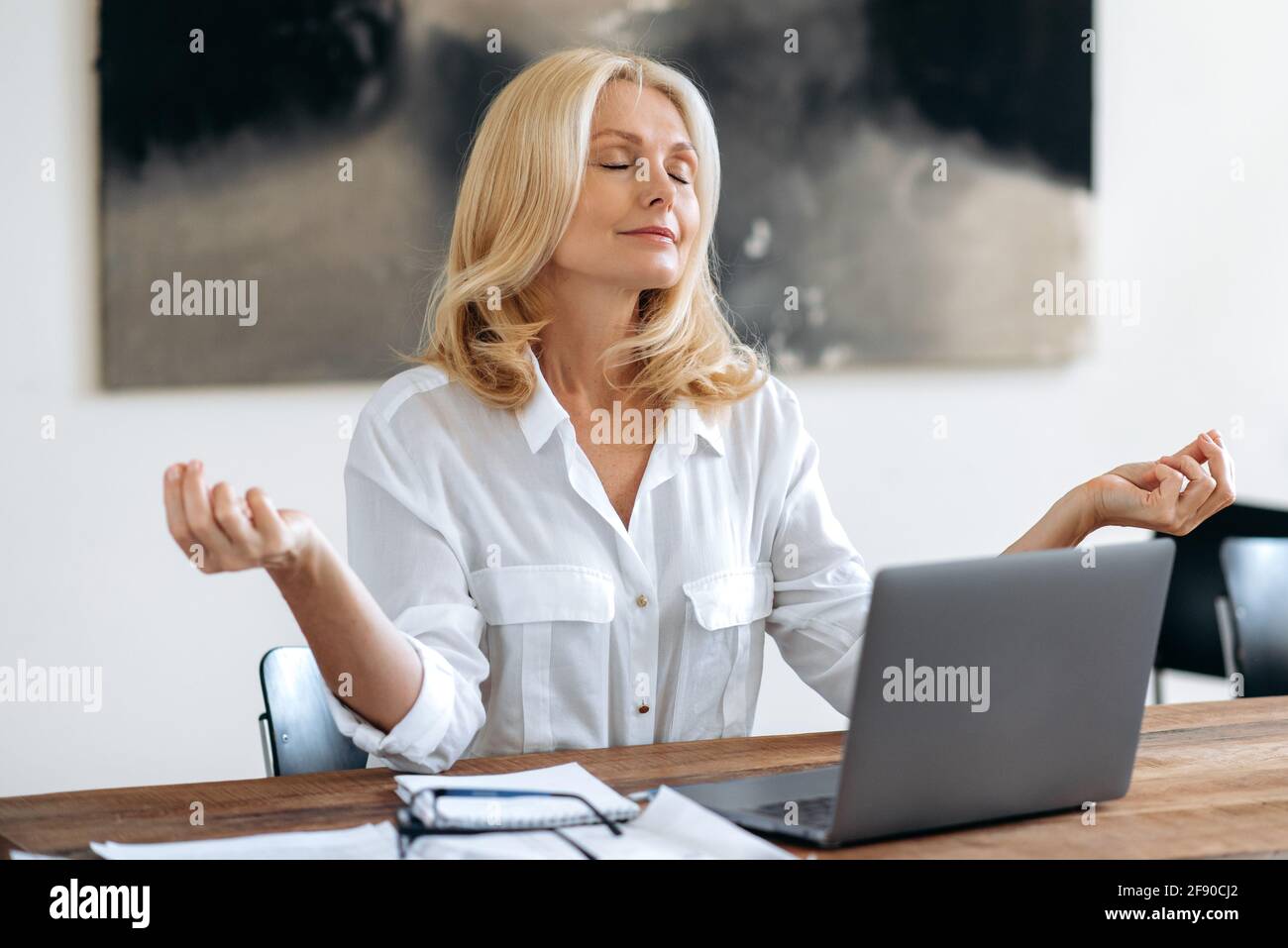 Schöne, erfolgreiche, entspannte, reife blonde Frau, Topmanagerin, Maklerin oder Freiberuflerin, in einem weißen Hemd, die am Laptop arbeitet, sitzt an einem Schreibtisch, ruht sich in einer Meditationhaltung aus, die Augen geschlossen, das Konzept der Ruhe Stockfoto