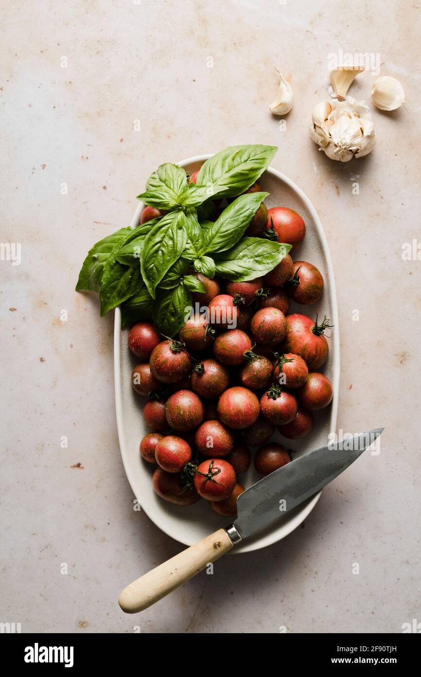 Ganze und in Scheiben geschnittene Kirschtomaten in einer Schüssel mit Basilikum- und Knoblauchzehen. Stockfoto