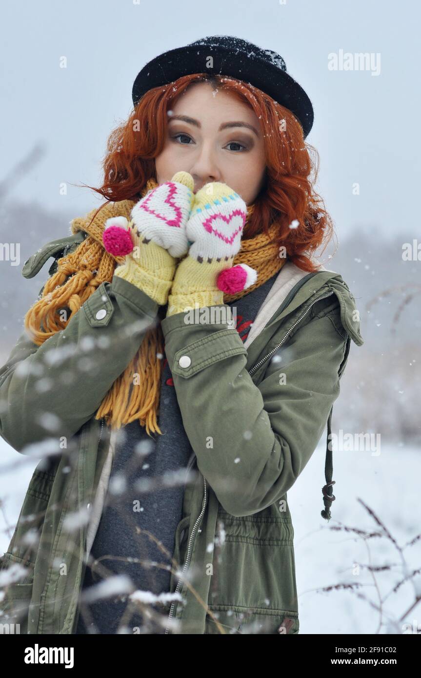 Rothaarige fröhliche Mädchen wärmt ihre Hände in warmen gestrickten Fäustlingen Im Feld bei Schneefall Stockfoto