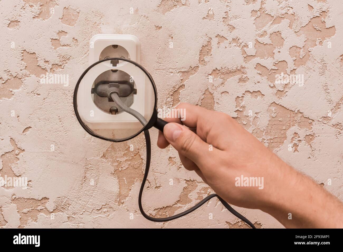 Die Hand eines Arbeiters mit einer Lupe untersucht ein Kabel, das an eine Doppelsteckdose angeschlossen ist. Das Konzept der Sicherheit von Haushaltsgeräten. Stockfoto