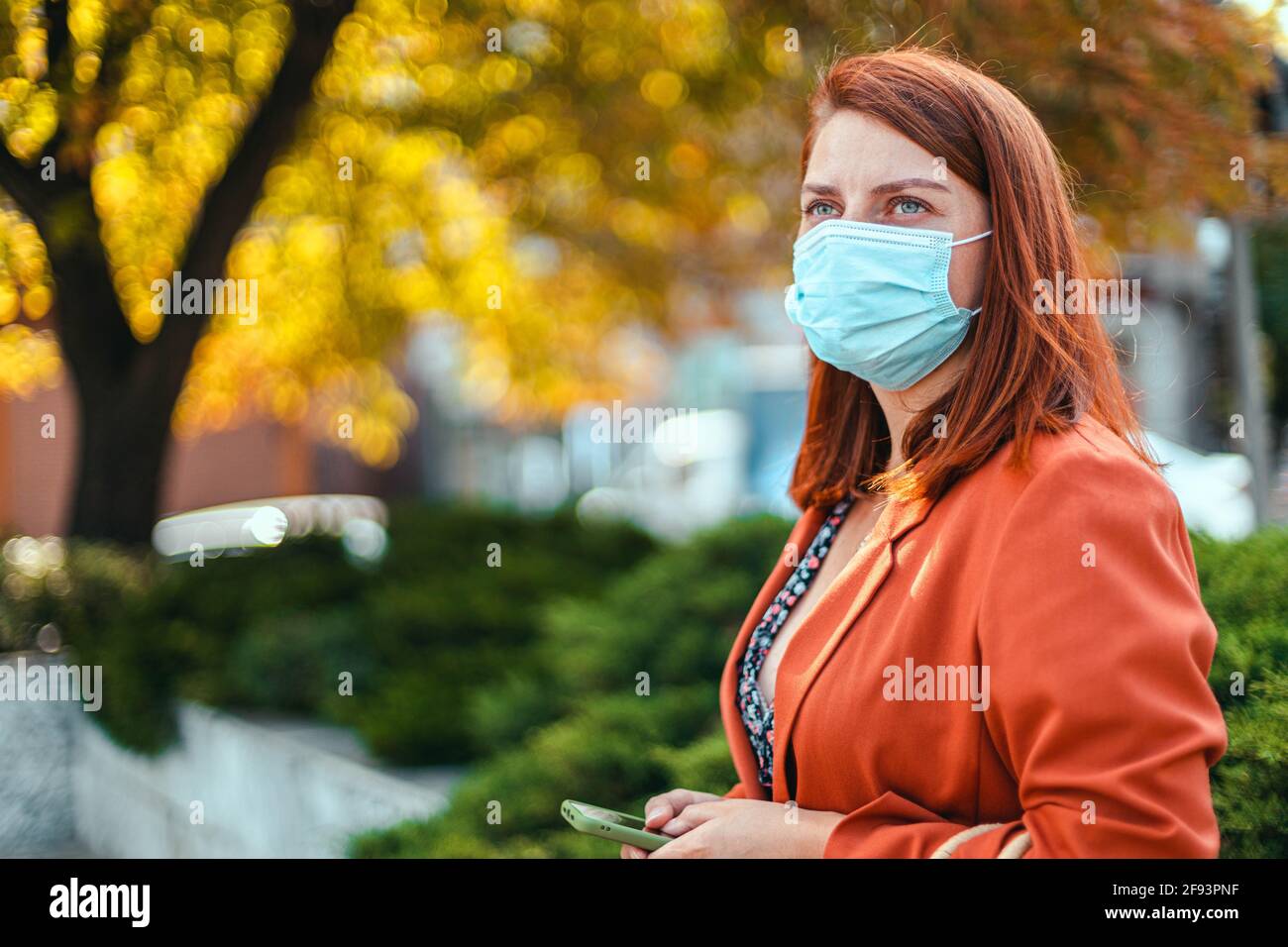 Geschäftsfrau mit Sicherheitsmaske, die in der Stadt steht. Schönes junges Mädchen mit roten Haaren in einer schützenden medizinischen Maske gegen allegria, Virus Stockfoto