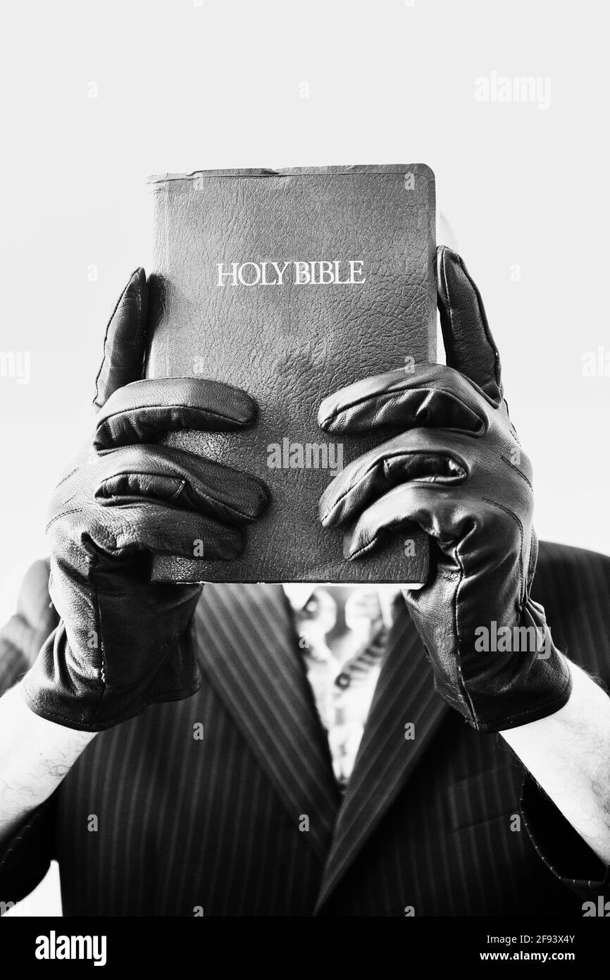 Mann mit schwarzen Lederhandschuhen und Nadelstreifenanzug, der die bibel hochhält, um sein Gesicht auf weißem Hintergrund zu bedecken. Konzept von Intrigen, Geheimnis, Neugier Stockfoto