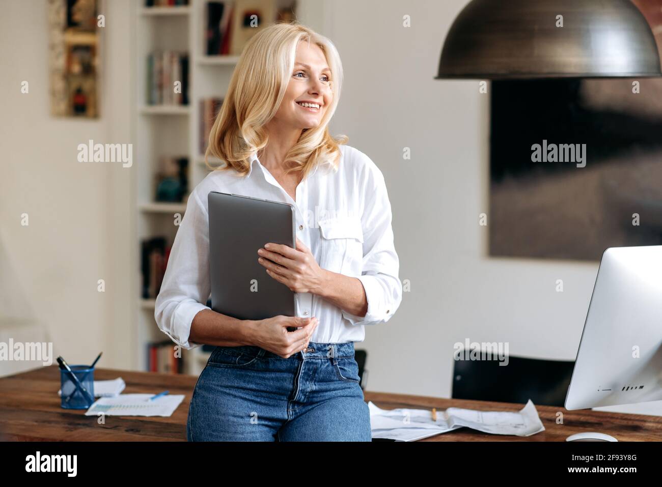 Porträt einer charmanten, eleganten kaukasischen blonden Frau, die in einem Büro in der Nähe ihres Arbeitsplatzes steht, in stilvoller Kleidung gekleidet, mit einem Laptop in der Hand, zur Seite schauend und freundlich lächelnd Stockfoto