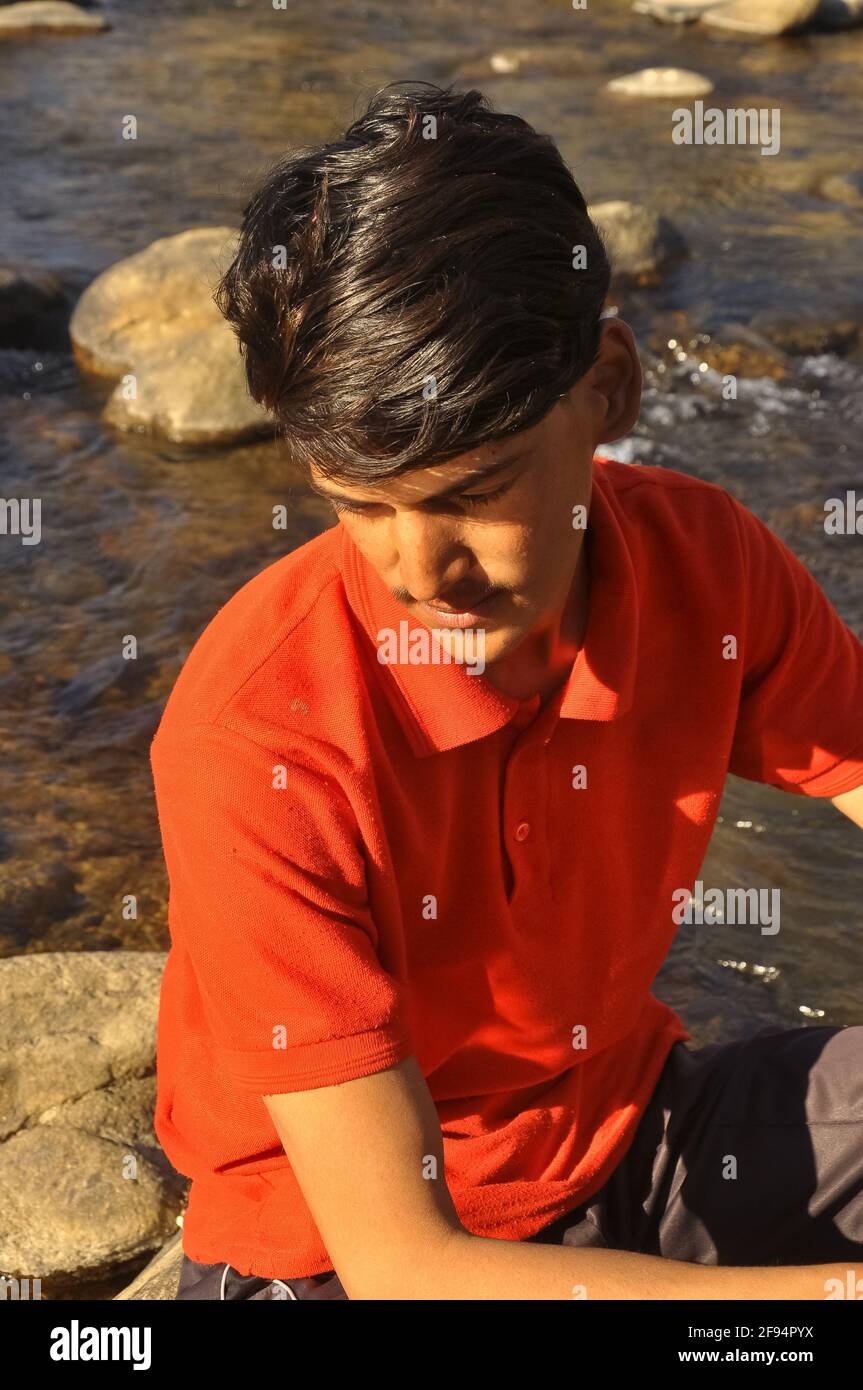 Ein junger Mann, der nach unten schaut und auf Stein sitzt, der neben dem Fluss liegt, junger Mann mit rotem T-Shirt. Stockfoto