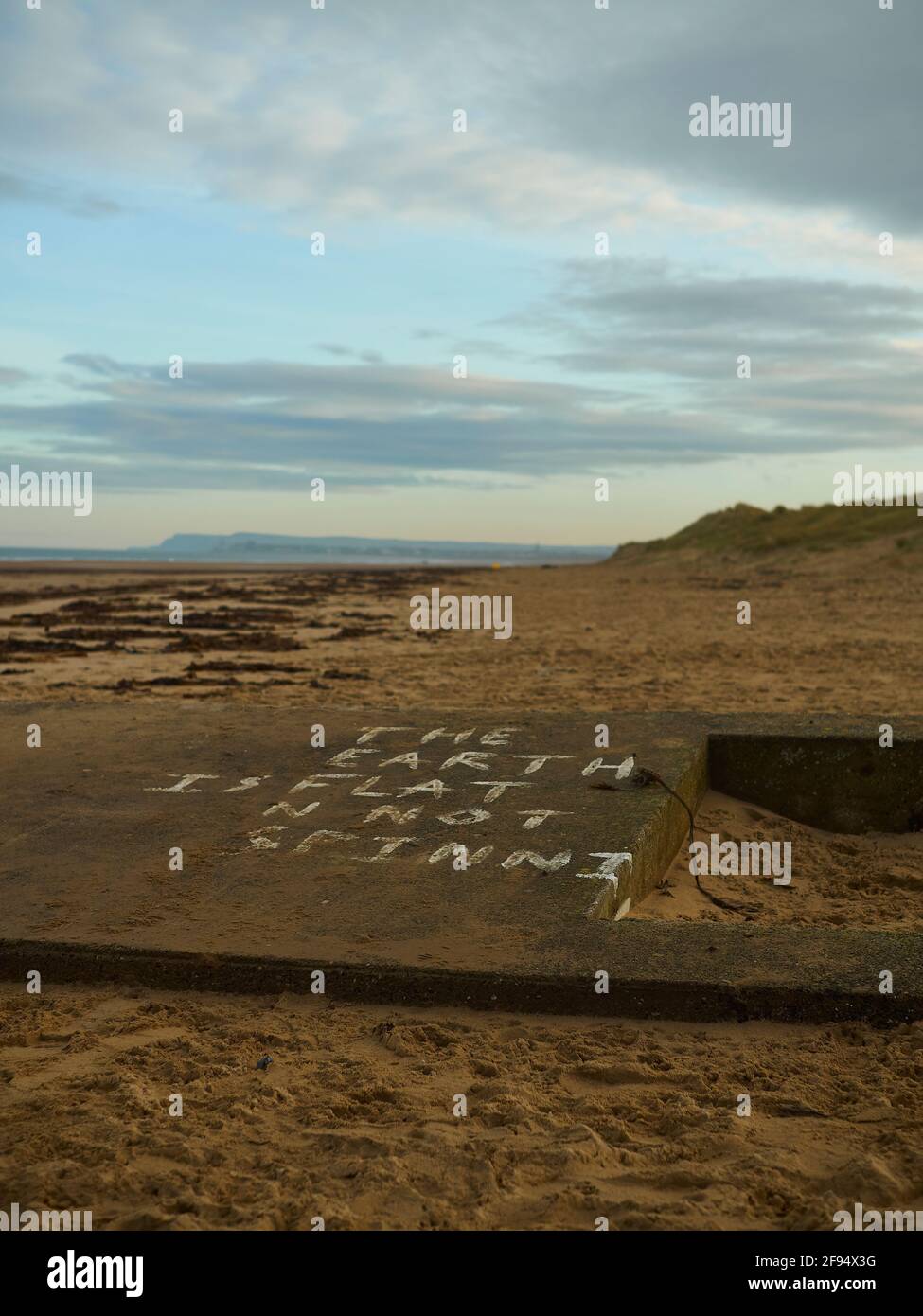 Ein Anti-Wissenschaft-/Flacherde-Slogan, der auf einem teilvergrabenen Bunker aus dem 2. Weltkrieg in den Sanddünen des Südbahnhofes bettet wird und am Strand entlang bis zu Hunt Cliff blickt. Stockfoto