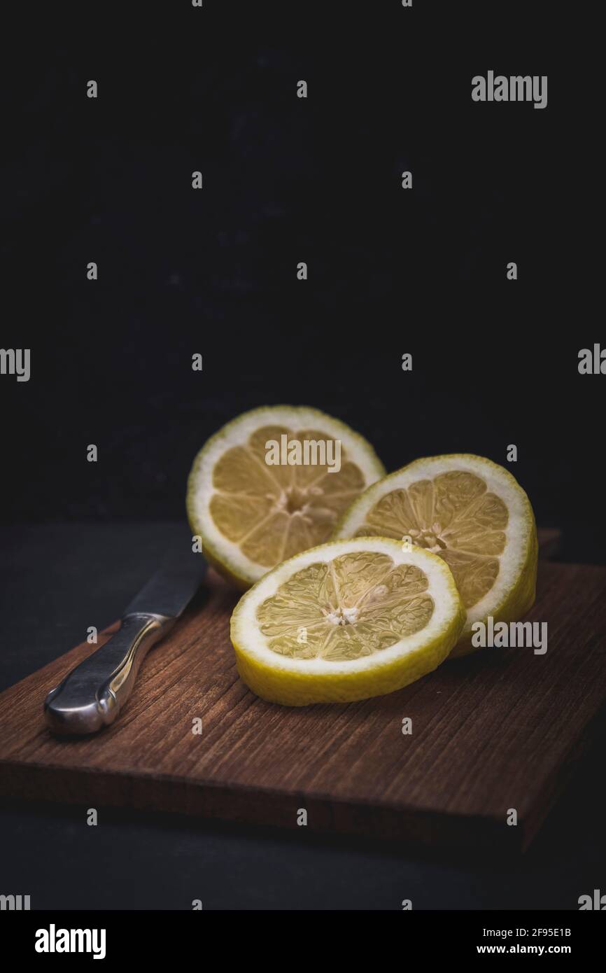 Zitrone in drei Stücke geschnitten, mit einem Messer auf einer Holzlatte liegend, vertikal mit Kopierraum und schwarzem Hintergrund Stockfoto