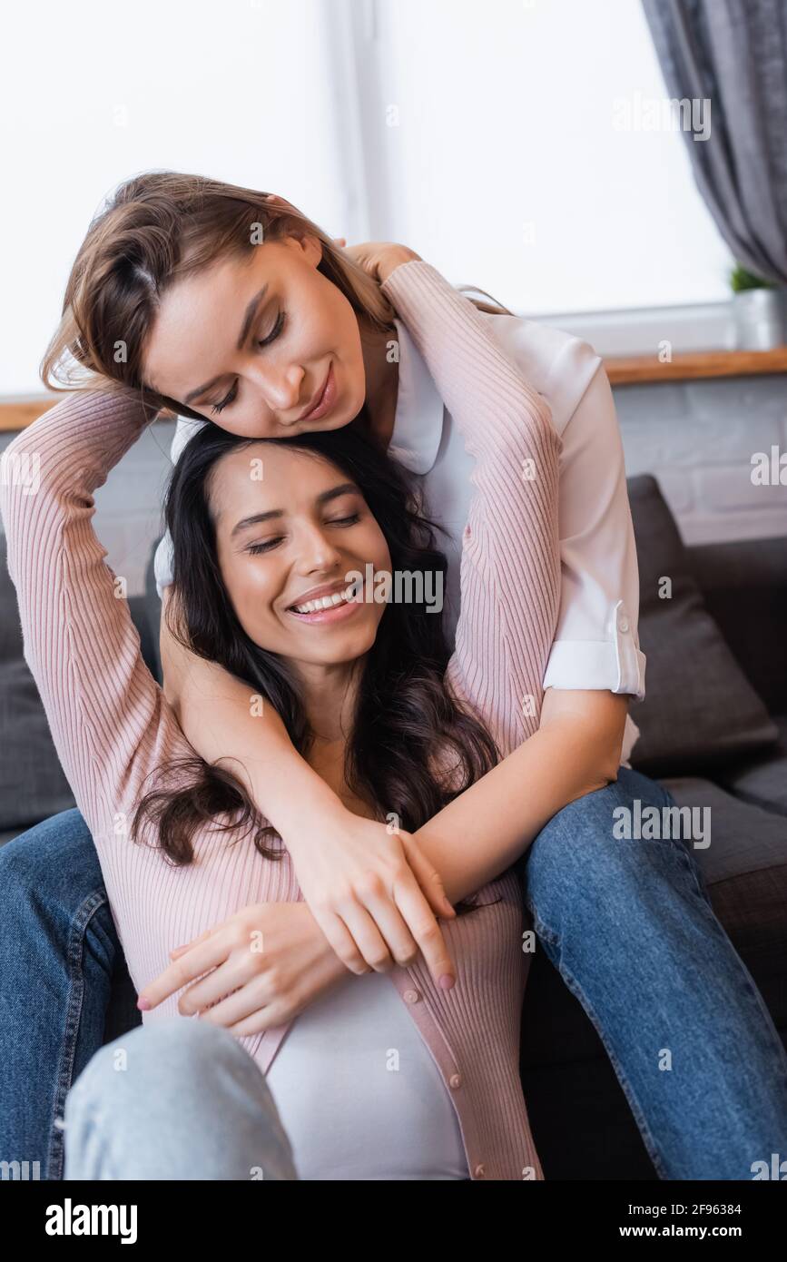Lächelnde Lesben mit geschlossenen Augen umarmen sich im Wohnzimmer Stockfoto
