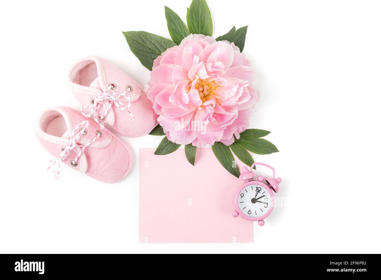 Pinke Babyschuhe mit Pfingstrose, Wecker und einer leeren Karte für die Nachricht auf weißem Hintergrund. Grußkarte oder Einladung für Neugeborene. Kopieren Sie den SPAC Stockfoto