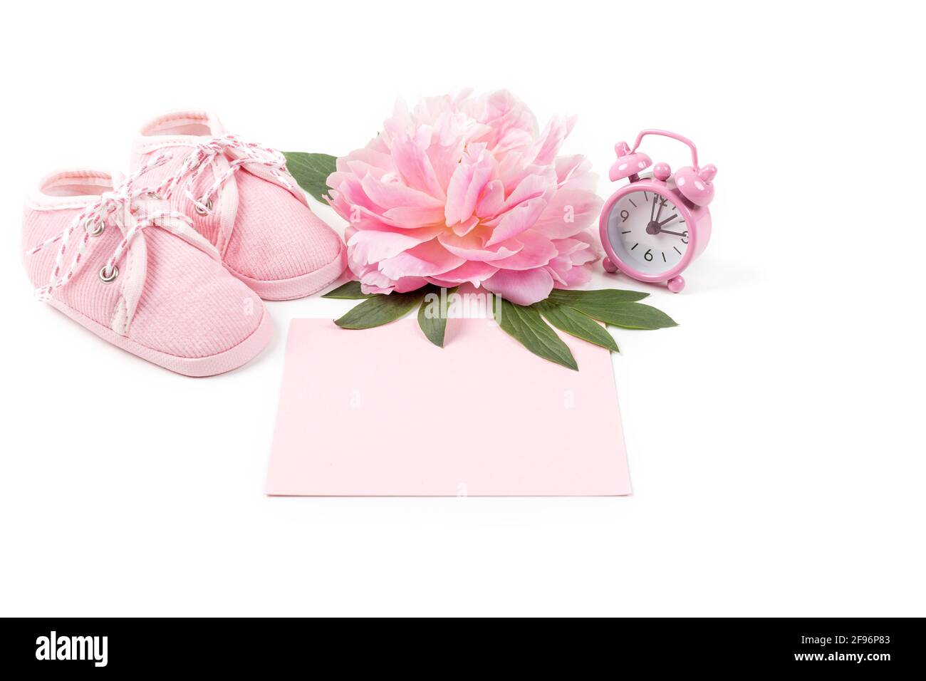 Pinke Babyschuhe mit Pfingstrose, Wecker und einer leeren Karte für die Nachricht auf weißem Hintergrund. Grußkarte oder Einladung für Neugeborene. Kopieren Sie den SPAC Stockfoto