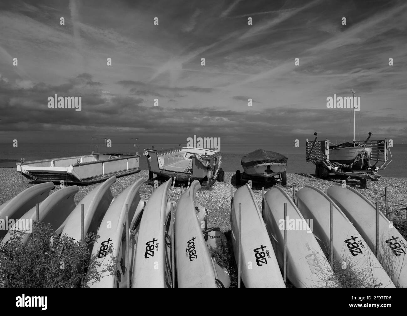 Monochromes Bild von Booten am Strand von Whitstable, Kent, Großbritannien, mit Flugzeugspuren am Himmel. Stockfoto