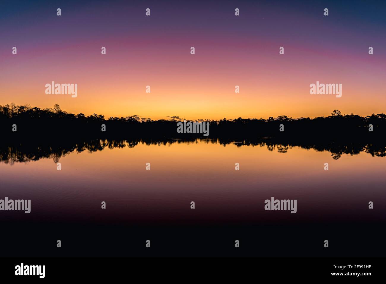 Ein Sonnenuntergang in der Abenddämmerung, der über einem stillen See mit einem Silhouettenhintergrund reflektiert wird. Es war ein schöner Nachmittag mit einem gelb-violetten Himmel Stockfoto