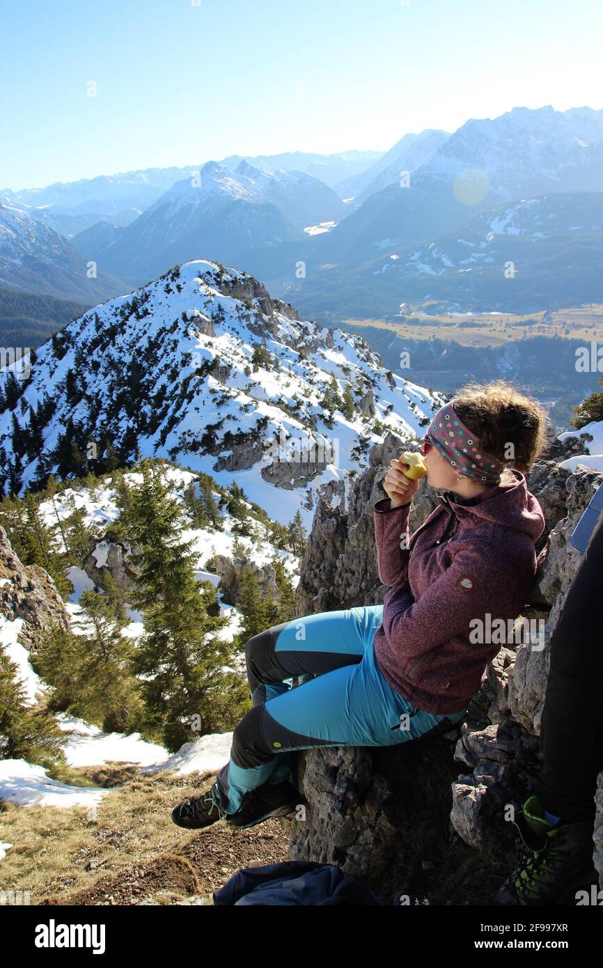 Junge Frau, die eine Pause macht, einen Apfel isst, Winterwanderung zum Signalkopf (1895 Meter) mit Blick auf den Wettersteingebirge, Zugspitze, Europa, Deutschland, Bayern, Oberbayern, Isartal, Krün Stockfoto