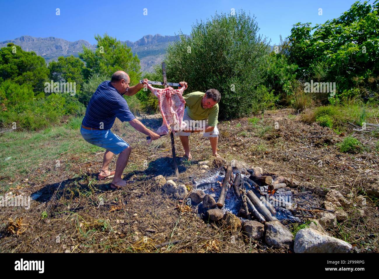 In Altea La Vella, Alicante, Spanien, grillen zwei Männer auf einem Eisenkreuz neben dem offenen Feuer Stockfoto