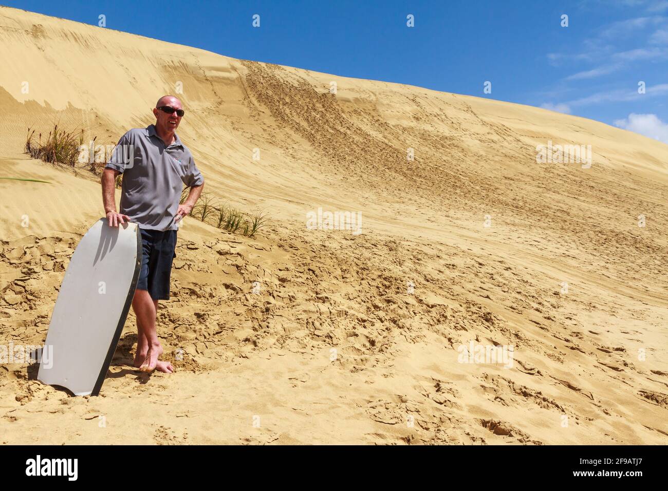 Ein Reiseleiter steht vor der riesigen Sanddüne in Te Paki, Northland, Neuseeland. Er hält ein Sandbrett, mit dem man die Düne hinunter reiten kann Stockfoto