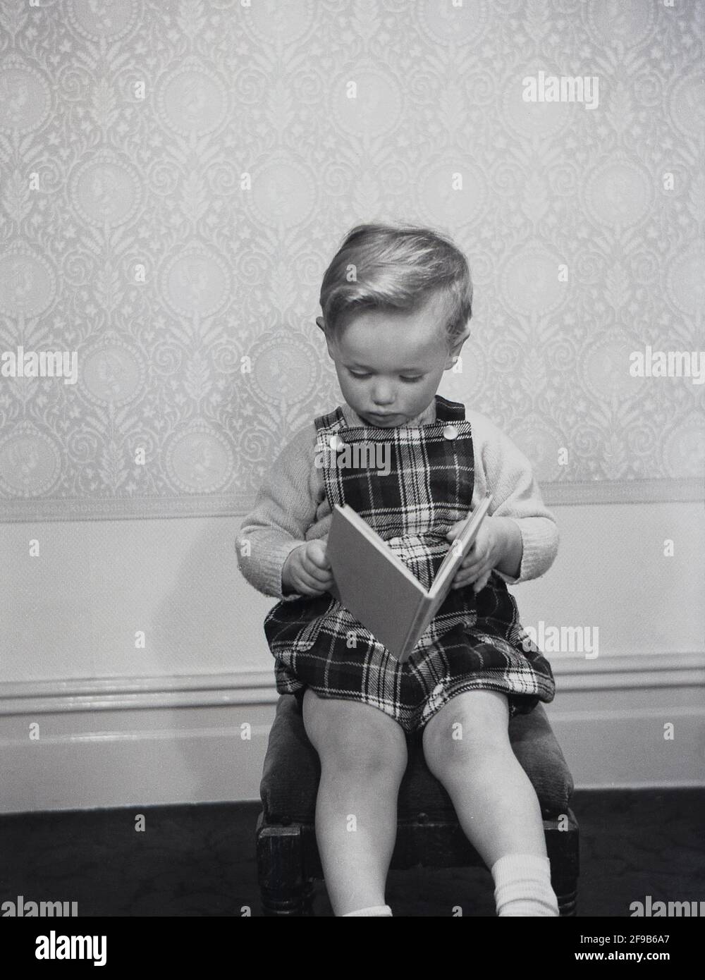 In den 1960er Jahren, historisch, in einem Raum, sitzt ein kleiner Junge mit einem Paar Tartan-gemusterter Latzhose auf einem Hocker und liest ein Buch, England, Großbritannien. Stockfoto