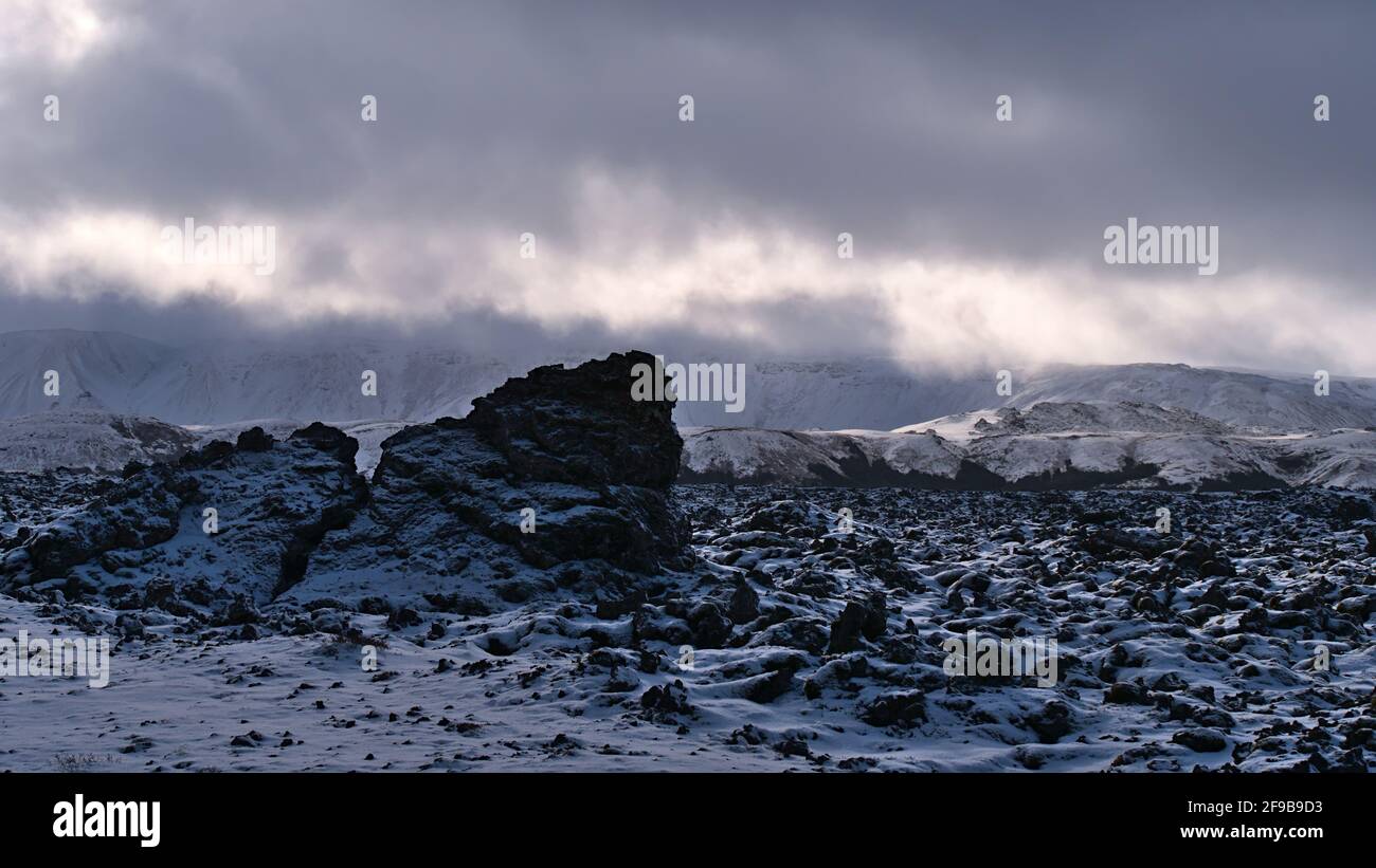 Wunderschöne Winterlandschaft mit schneebedeckten Lavafeldern mit vulkanischen Felsen und schroffen Bergen im Hintergrund mit Wolken und Sonnenlicht in Island. Stockfoto