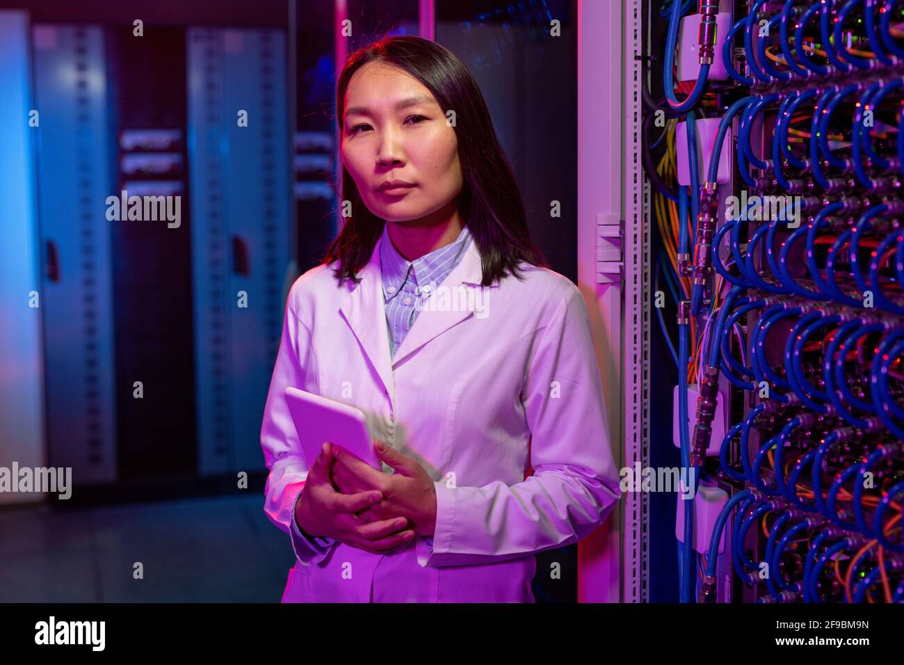 Porträt eines attraktiven asiatischen Serverprofi in weißem Mantel Stehend mit Tablet am offenen Serverschrank mit angeschlossenen Kabeln Stockfoto