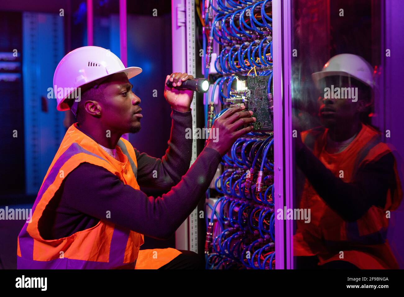 Konzentrierter schwarzer Arbeiter in Hardhut und orangefarbener Weste, der sich verduckt Server und Verwendung von Taschenlampe bei der Behebung des Serverproblems Stockfoto