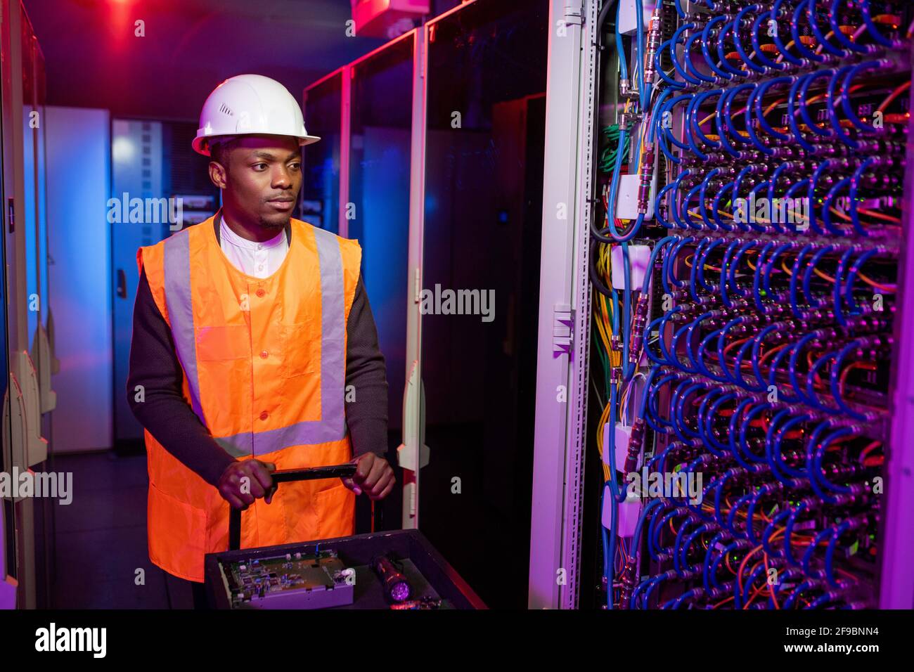 Seriöser schwarzer Elektriker im Rechenzentrum in einem Hardhut, der sich mit dem Wagen abschob Tools während der kommenden Serverschrank zur Reparatur Stockfoto