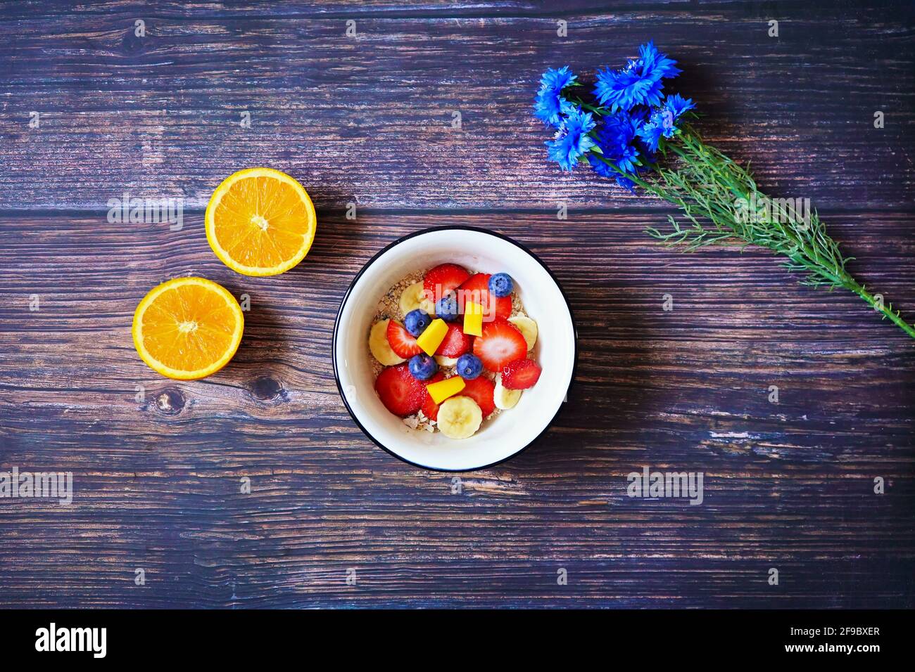 Gesunde Frühstückschale mit Cerealien und frischen Früchten (Erdbeere, Banane, Heidelbeere, Mango) und Orangenfrüchten auf einem hölzernen Frühstückstisch. Stockfoto