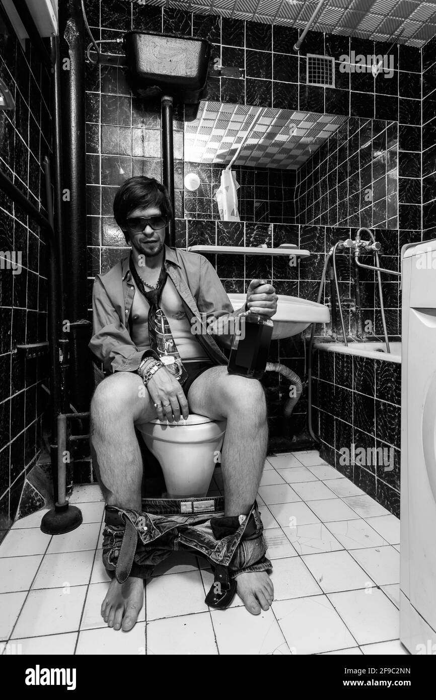 Betrunkener Mann sitzt in einer Toilette mit einer Alkoholflasche Stockfoto