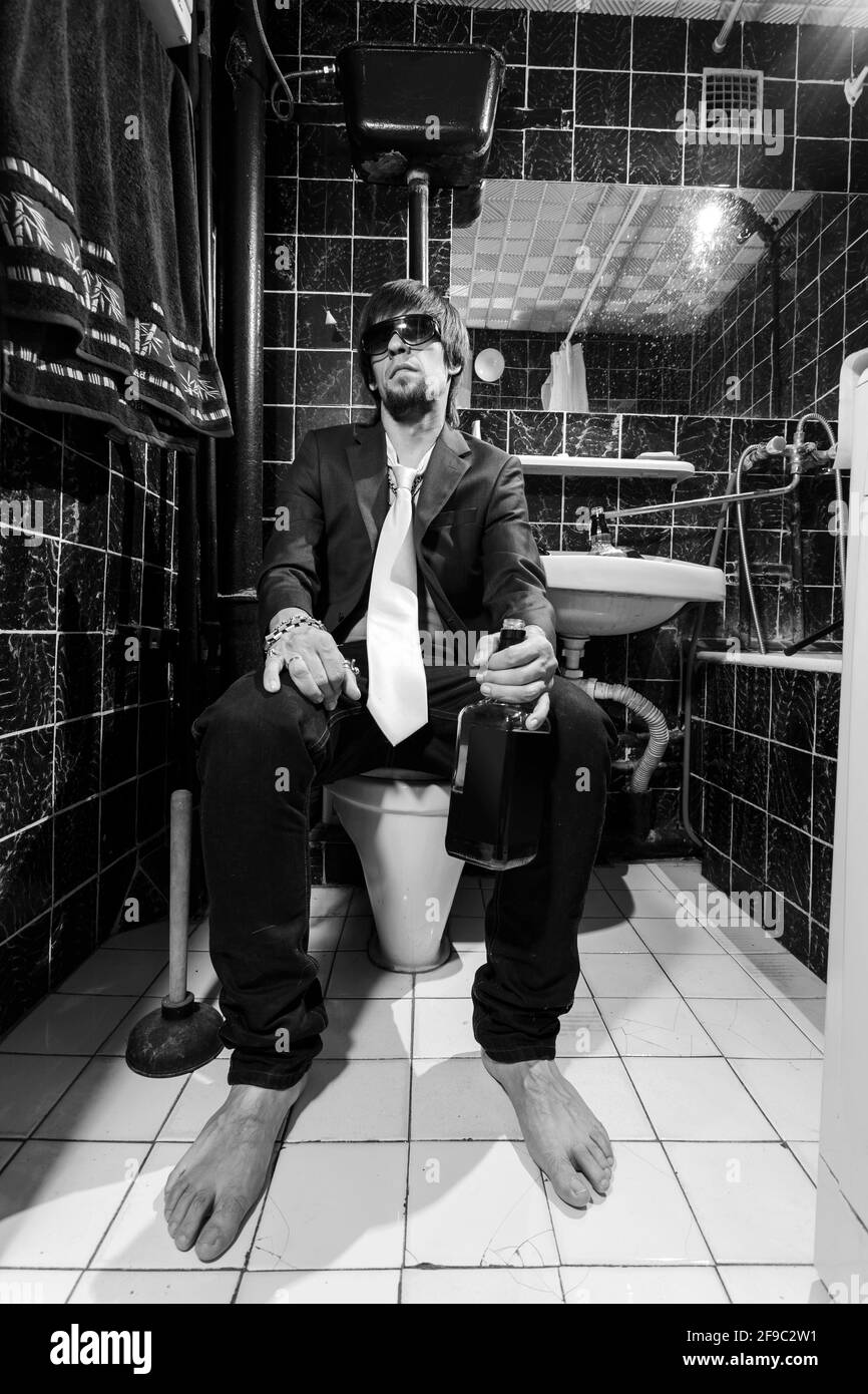 Betrunkener Mann sitzt in einer Toilette mit einer Flasche Whiskey Stockfoto