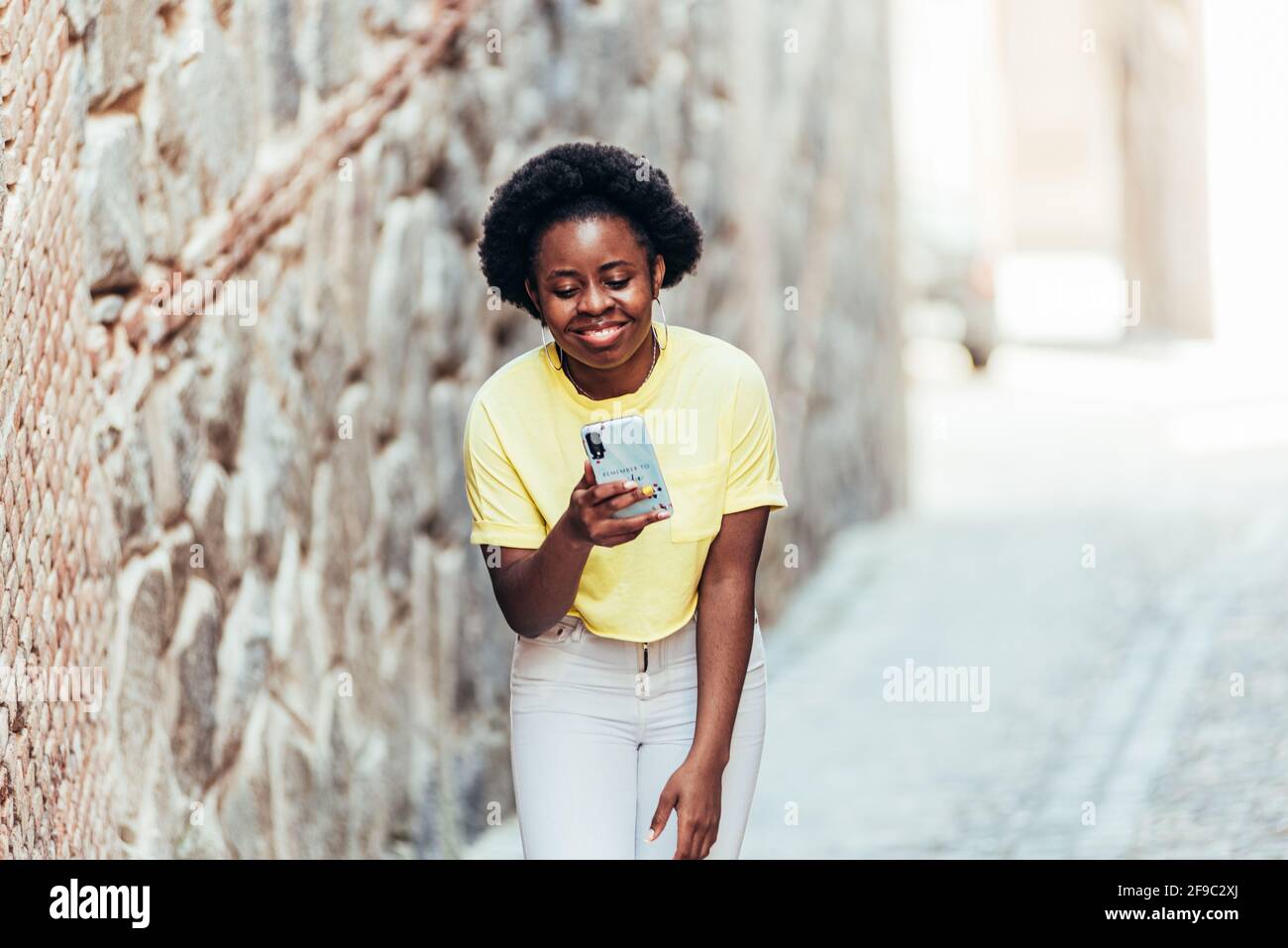 Porträt eines schwarzen afroamerikanischen Mädchens, das lächelt, während sie ihr Handy auf einer Straße in der Altstadt benutzt. Stockfoto