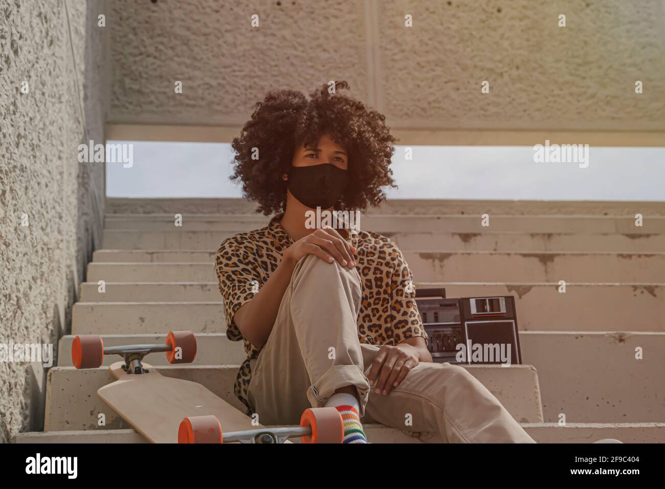 Junger Afro-Mann mit Maske sitzt auf einigen Treppen mit Ein Longborad Stockfoto