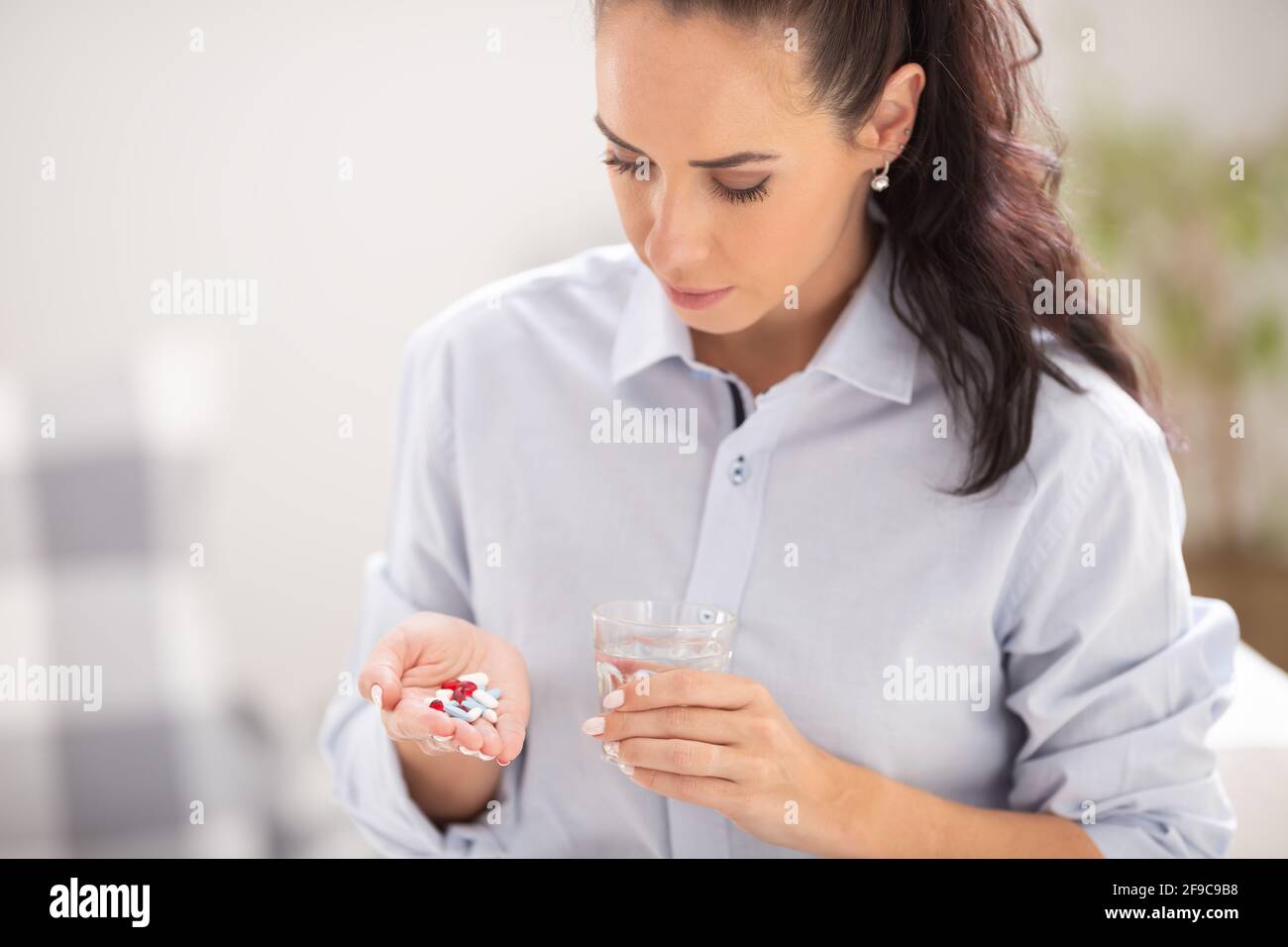 Gut aussehende Frau schaut auf die Hand mit vielen Medikamenten oder Nahrungsergänzungsmitteln, die planen, sie zusammen mit einem Glas Wasser zu nehmen. Stockfoto