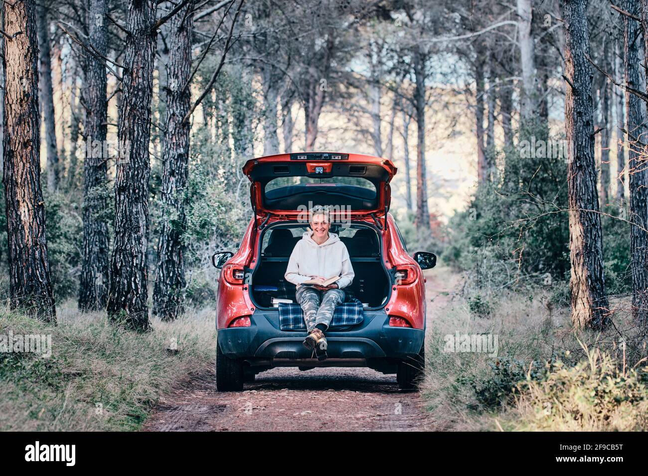 Junge Erwachsene kaukasische Frau liest ein Buch auf dem offenen Kofferraum eines Autos, das auf einem Waldweg geparkt ist. Reisekonzept. Stockfoto