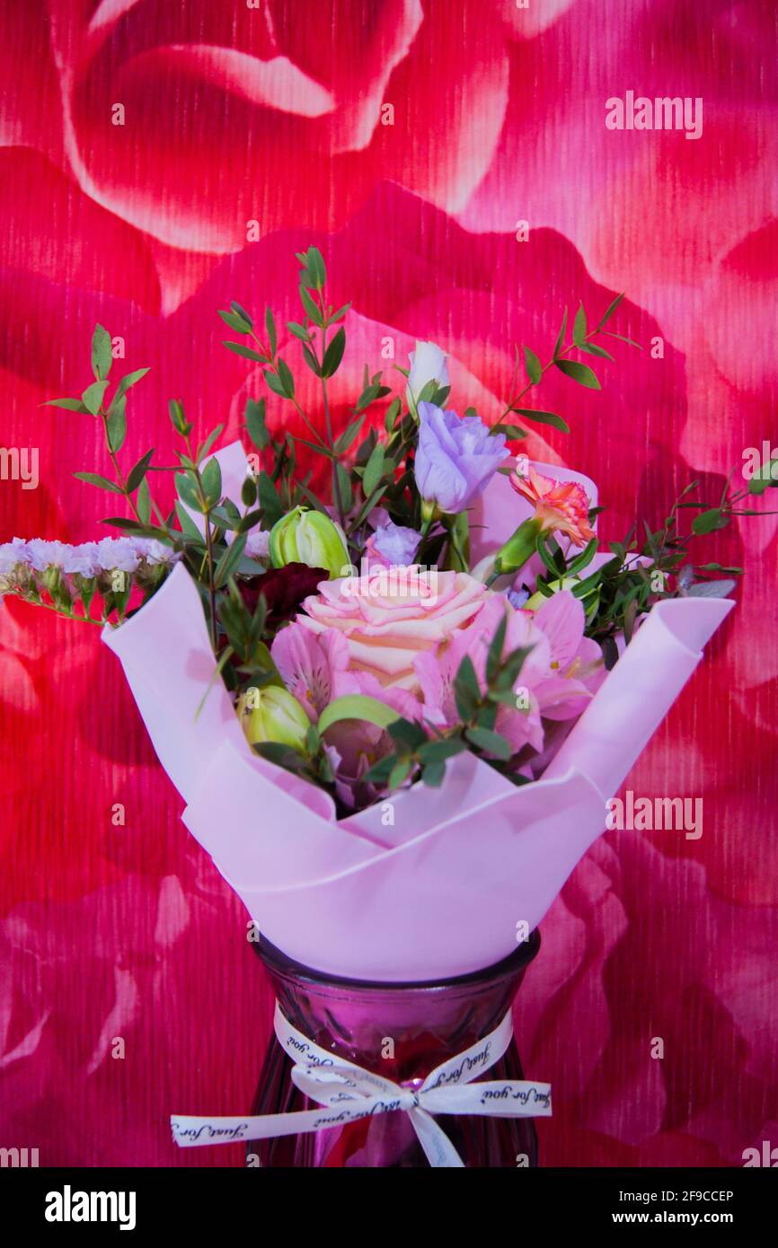 Ein zartes Blumenstrauß mit verschiedenen Blumen in einem hellrosa Wrapper steht in einer häuslichen Umgebung gegen eine bunte rosafarbene Wand. Vertikales Foto. Stockfoto
