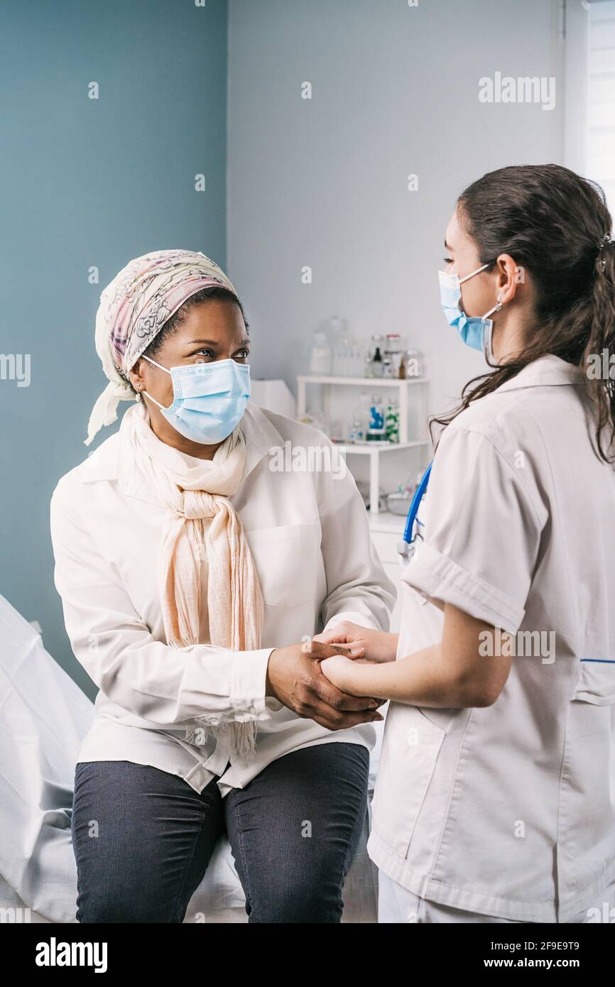 Junge Ärztin in medizinischer Uniform und Stethoskop trägt Gesichtsmaske sprechen und hält die Hände von afroamerikanischen reifen Frau Patientin während AP Stockfoto