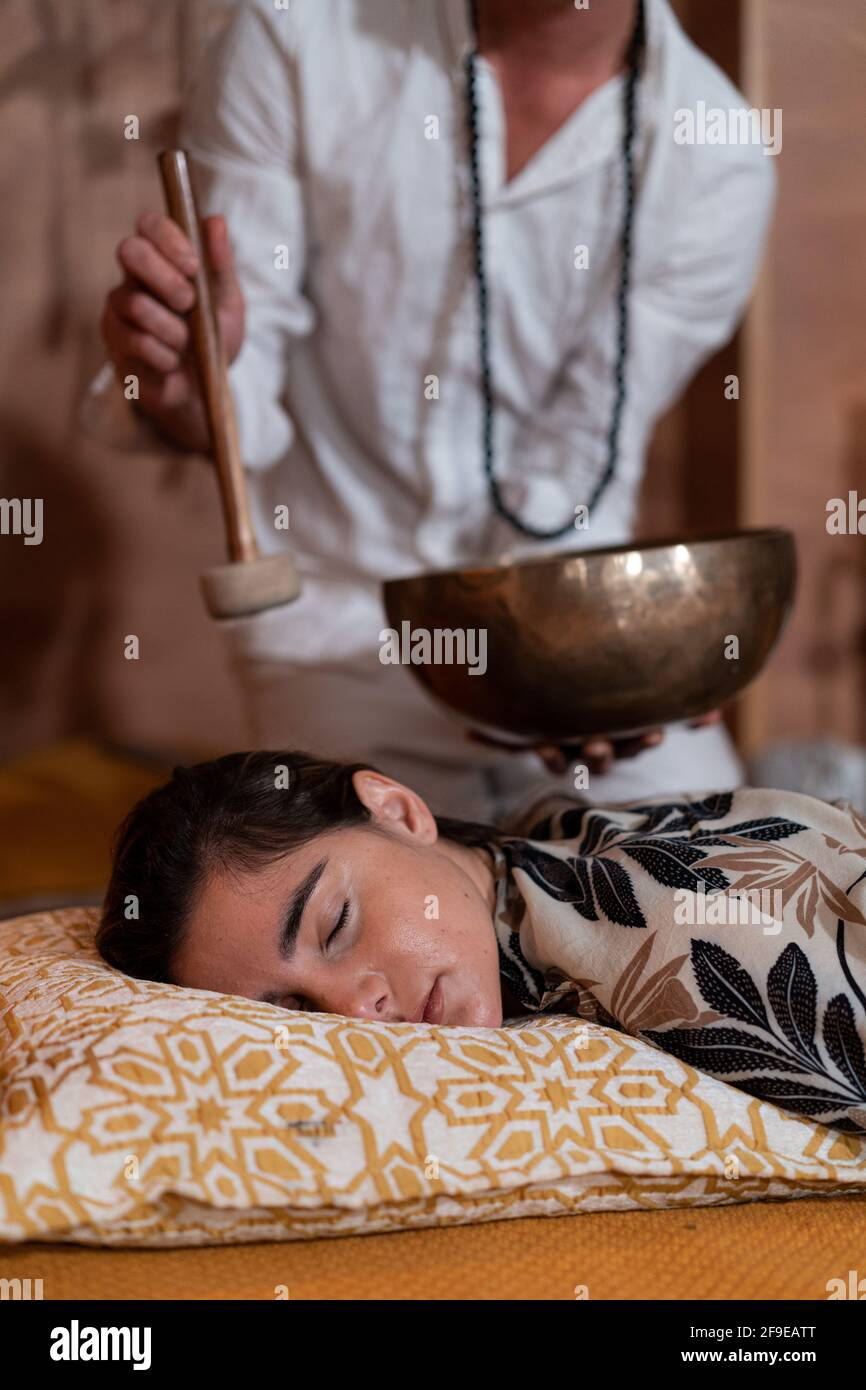 Zugeschnittenes, nicht erkennbarer männlicher spiritueller Therapeut in Perlen, die tibetischen Gesang spielen Schale über junges Weibchen mit geschlossenen Augen Stockfoto