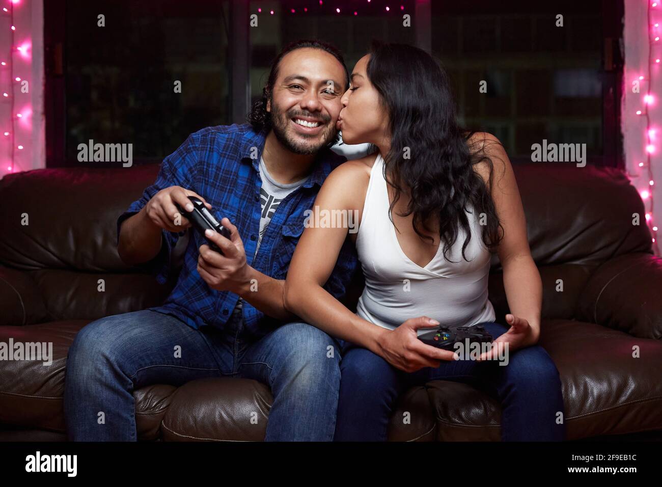 Aufgeregtes ethnisches Paar in Freizeitkleidung mit spielenden Joy Pads Videospiel zusammen, während Sie zu Hause auf der Ledercouch sitzen Stockfoto